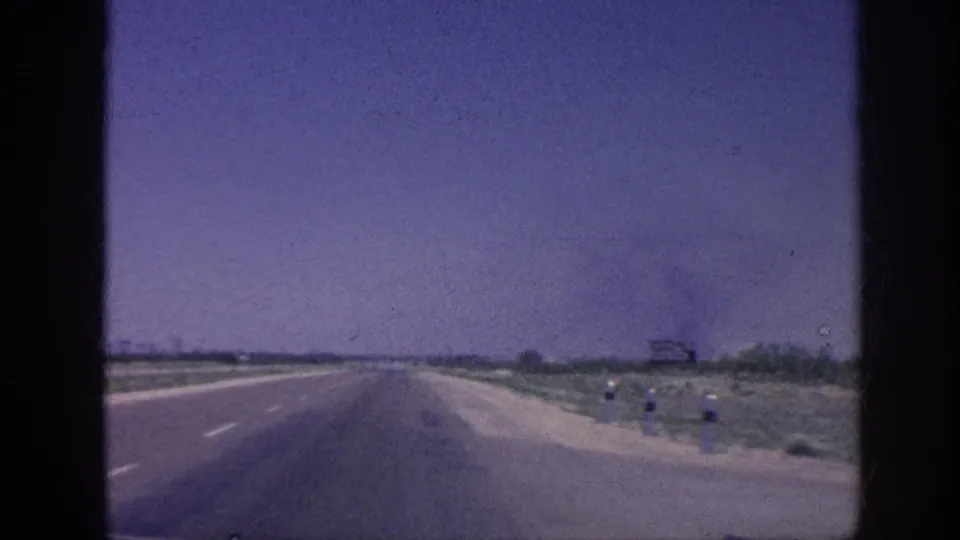 8mm windshield POV of a straight American highway, 1963, with a dark smoke plume on the left horizon, sparse roadside scrub, and a distant billboard at right.