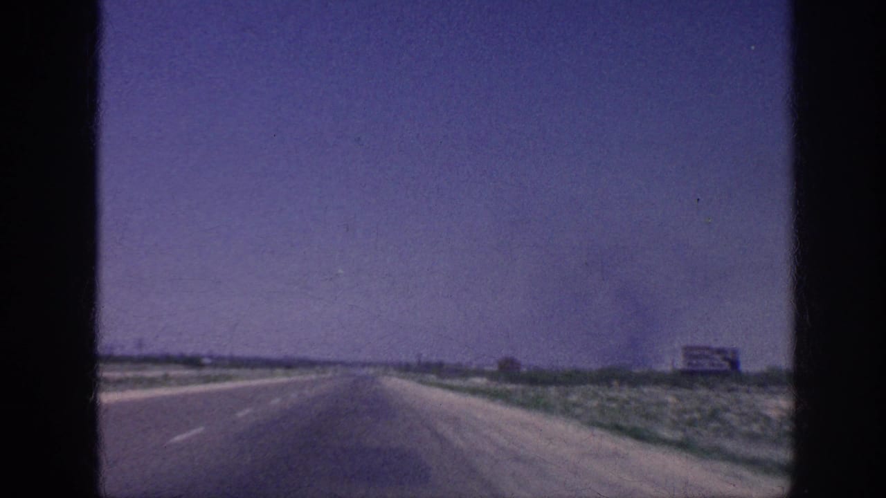 Straight divided highway stretches to the horizon under a blue-purple sky, with low scrubland on both sides and a distant billboard on the right shoulder.