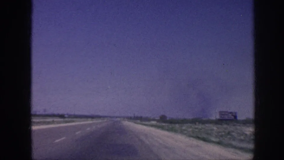 Windshield POV of an American highway, mid-1960s, with flat scrubland flanking the road and a large rectangular billboard structure visible in the far right distance.