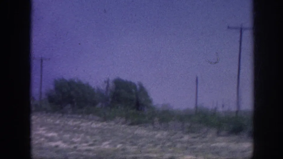8mm home movie frame from what appears to be a rural area near Memphis, Tennessee, 1963 — a car window view of dry grassland, trees, and utility poles against a purple-shifted sky.