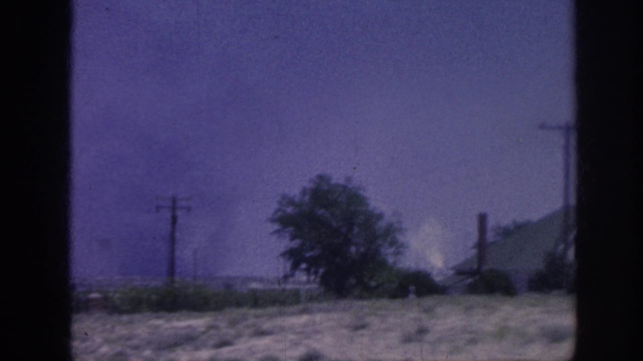 8mm home movie frame, c.1963, rural landscape shot through a window: trees, utility poles, distant smoke plume, purple-shifted sky.