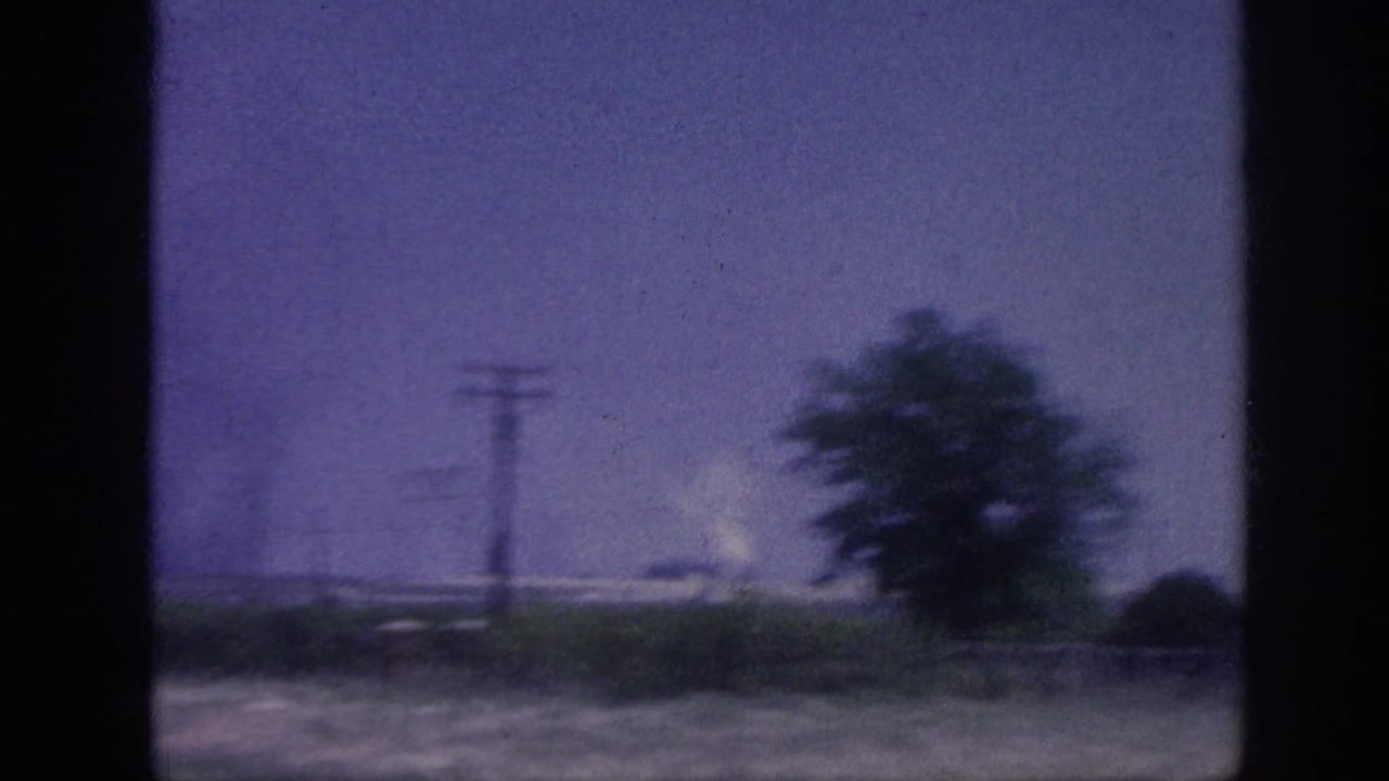 Rural field through window showing a tree, utility poles, and a distant white smoke plume against a purple sky.