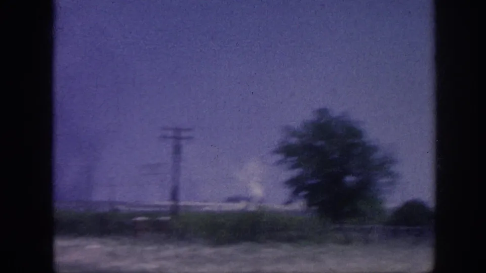 8mm home movie c.1963, rural Tennessee landscape through window: tree, utility poles, white smoke plume on horizon, aged purple color shift.