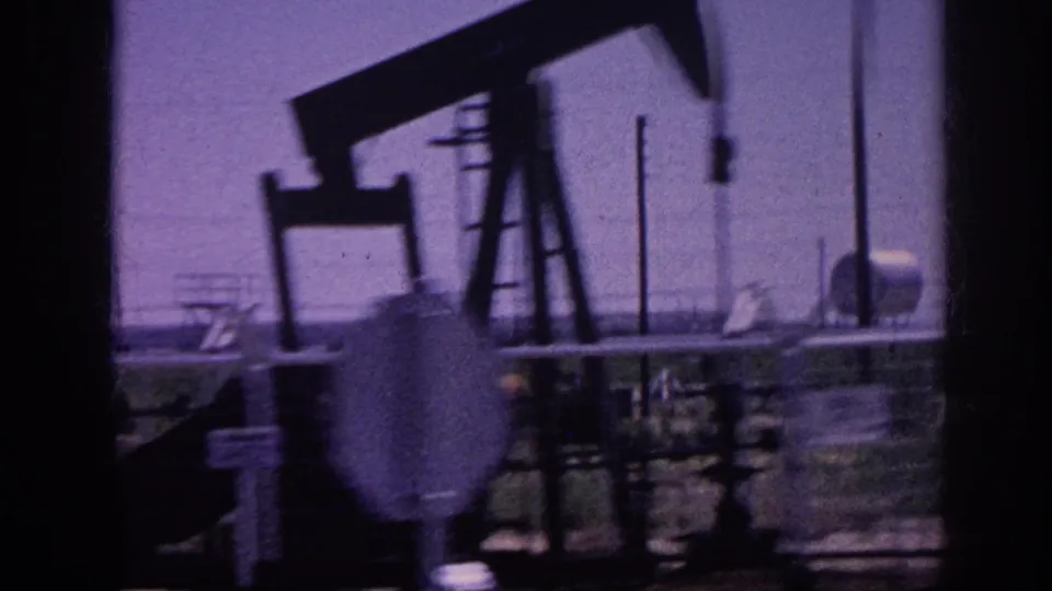 8mm home movie c.1963, oil pumpjack silhouetted against an aged purple sky, industrial storage structures and poles visible behind.
