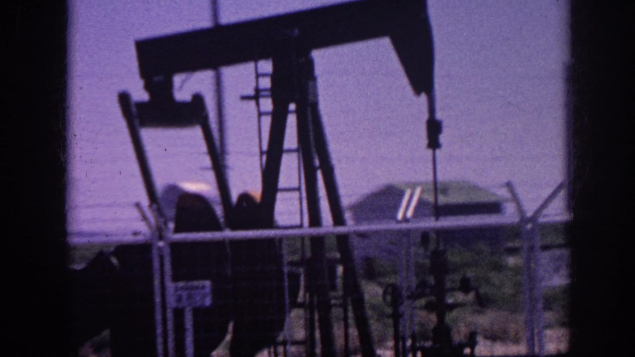 8mm home movie frame circa 1963 showing an oil pump jack in silhouette against a purple dye-shifted sky, shot from inside a moving car with a chain-link fence across the foreground.