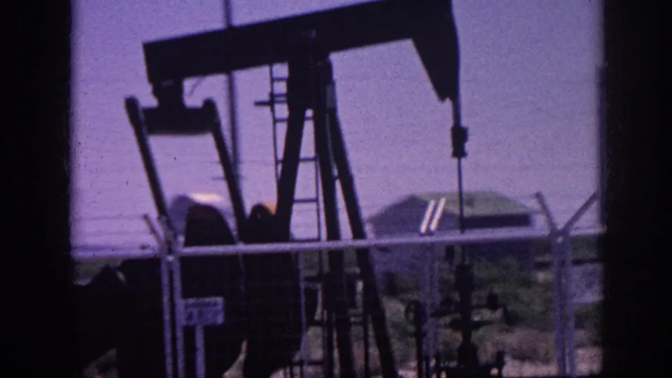 8mm home movie frame circa 1963 showing an oil pump jack in silhouette against a purple dye-shifted sky, shot from inside a moving car with a chain-link fence across the foreground.