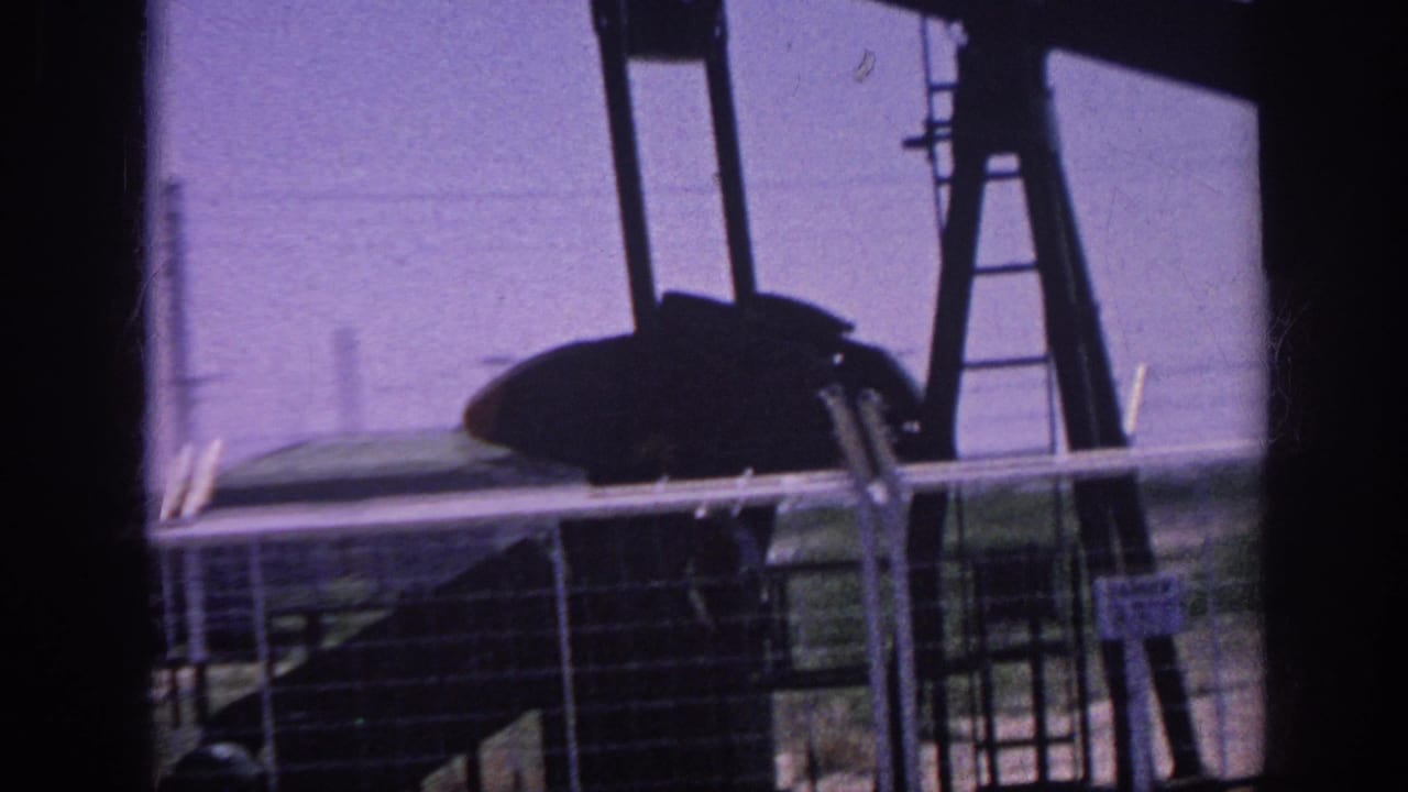 Oil pump jack silhouette and a dark-profiled passenger seen through a car window against a purple-tinted sky.