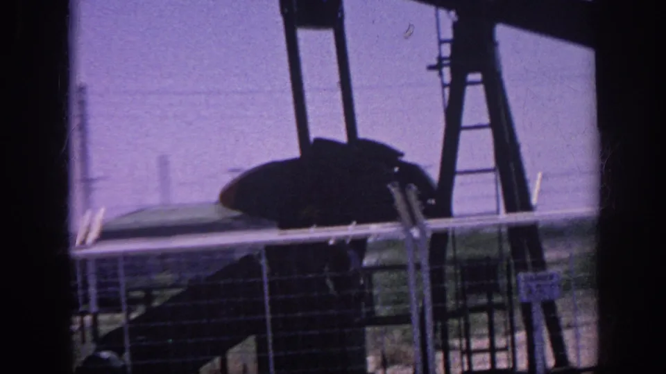 8mm film frame circa 1963 showing an oil pump jack and a silhouetted passenger in profile viewed from inside a moving car, with utility poles and chain-link fence visible.
