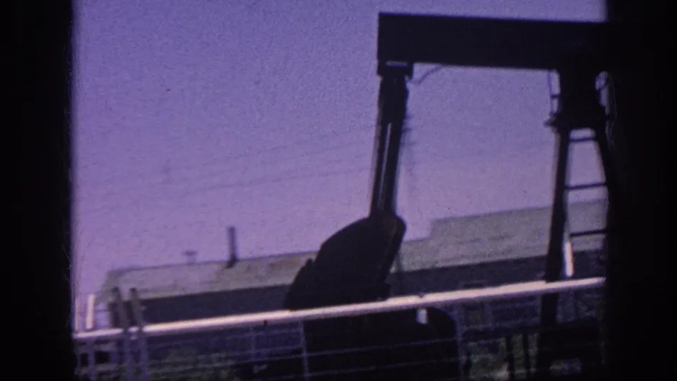 8mm home movie frame circa 1963 showing a close pump jack silhouette against a purple-tinted sky, framed through a moving car window with a chain-link fence in the foreground.