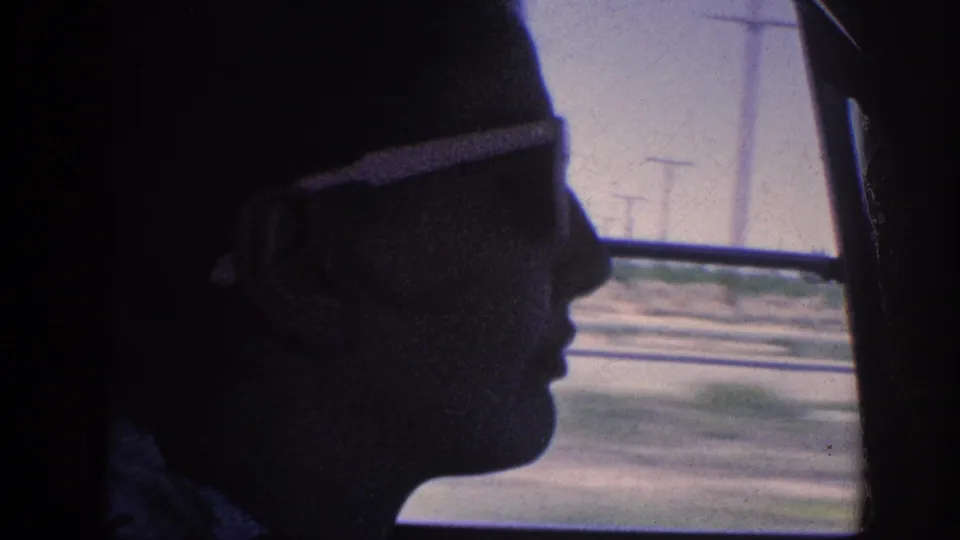 8mm home movie circa 1963 showing a silhouetted passenger in close profile wearing sunglasses, framed against a passing road and power lines through the car window.
