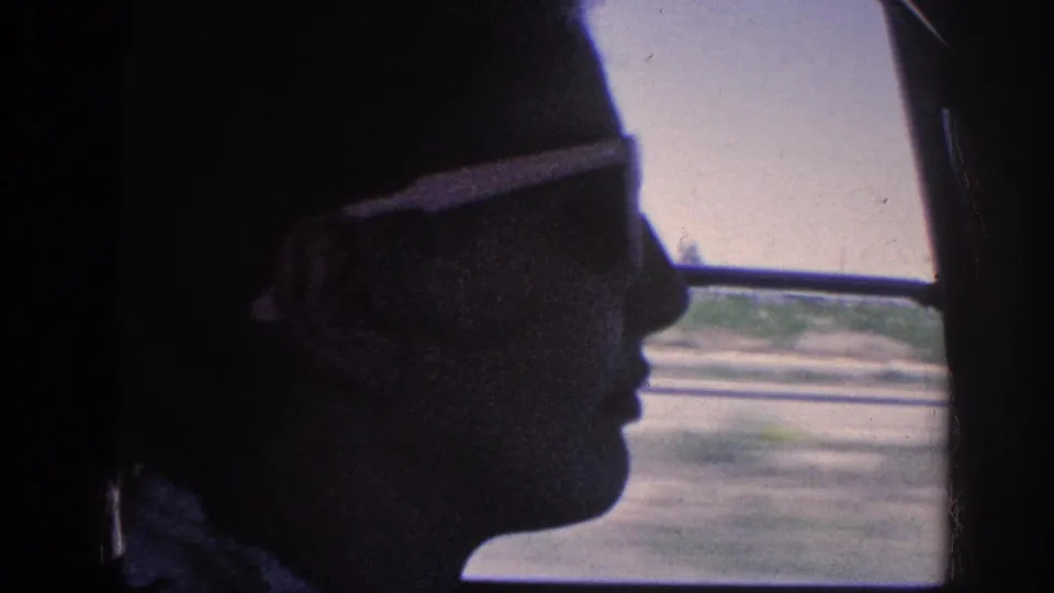 Silhouetted traveler with sunglasses in close profile inside a moving car, flat rural road and landscape visible through the window; Memphis, Tennessee, circa 1963.