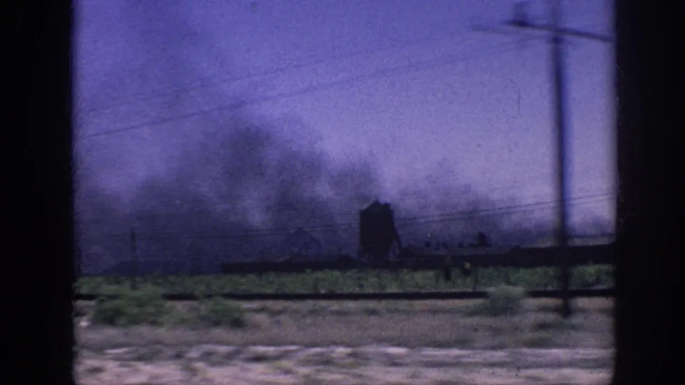 View through car window of flat rural terrain with dark smoke plumes rising in the distance, utility lines crossing the frame, green fields below; blue-purple color cast from aged 8mm stock, possibly near Memphis, 1963.