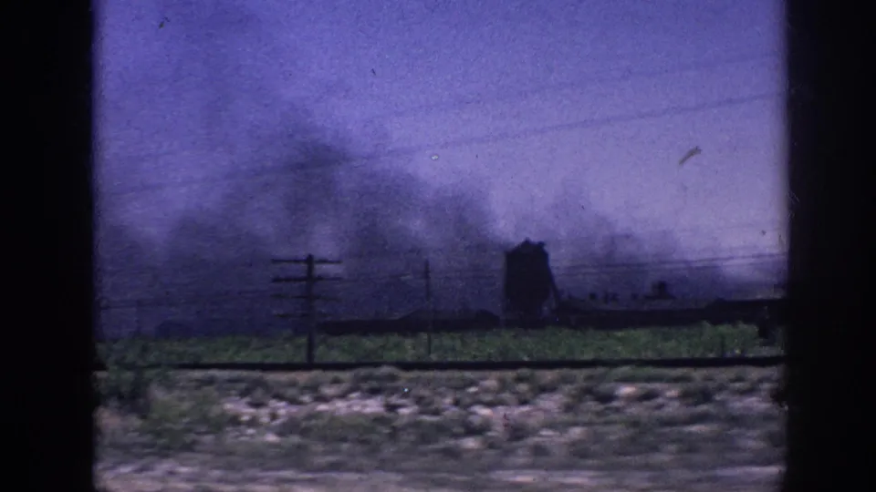 Flat rural terrain with dark smoke rising on the horizon, utility poles, and sparse vegetation seen through a car window; deep purple color cast from aged 8mm stock, possibly near Memphis, 1963.