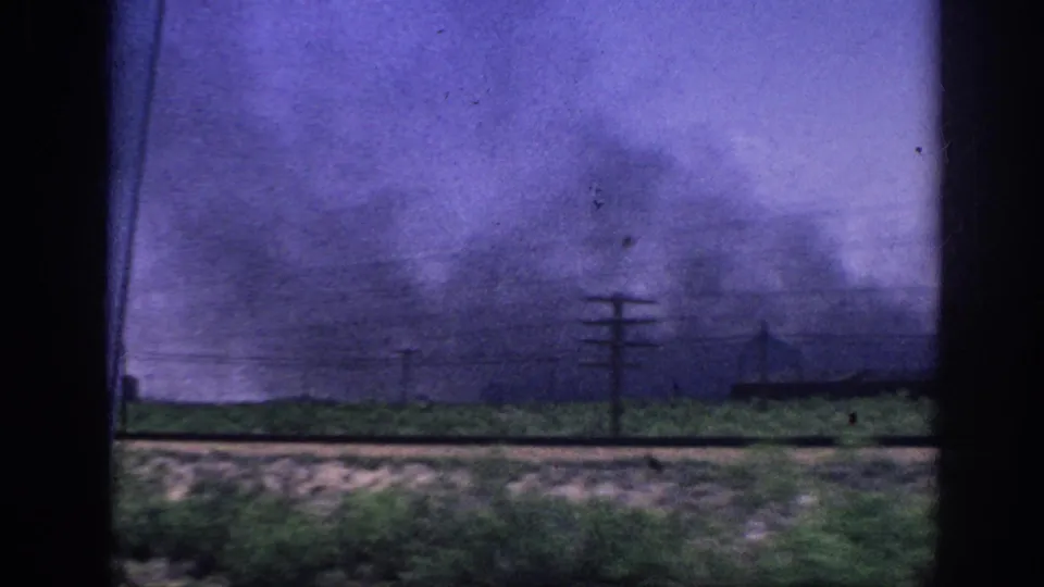 8mm home movie, c.1963 Memphis — a broad diffuse smoke plume spreads over flat terrain with multiple utility poles in silhouette, shot from a moving vehicle window.