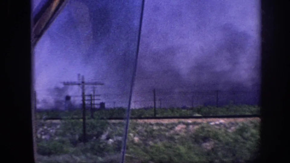 8mm home movie, c.1963 Memphis — smoke over flat terrain with utility poles and a faint mid-ground structure; vehicle window frame visible at upper right.