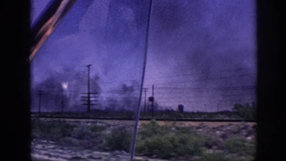 1963 Memphis, Tennessee area: view through a moving vehicle window of a roadside with utility poles and large dark smoke plumes under a color-shifted purple sky.