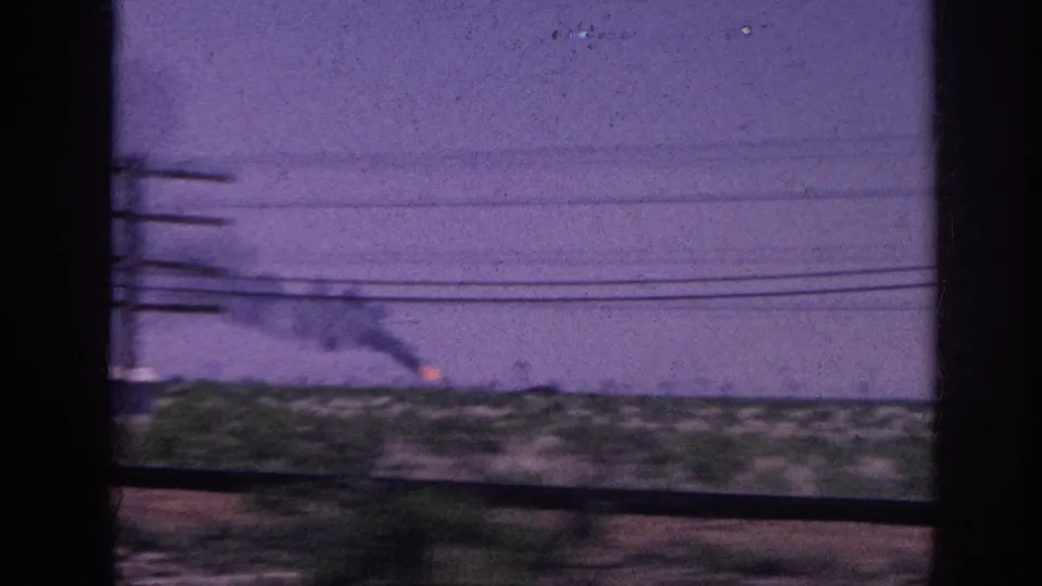 1963 Memphis, Tennessee area: vehicle window view of an open flat rural landscape with power lines crossing the frame and a small fire with smoke at the left under a hazy purple sky.