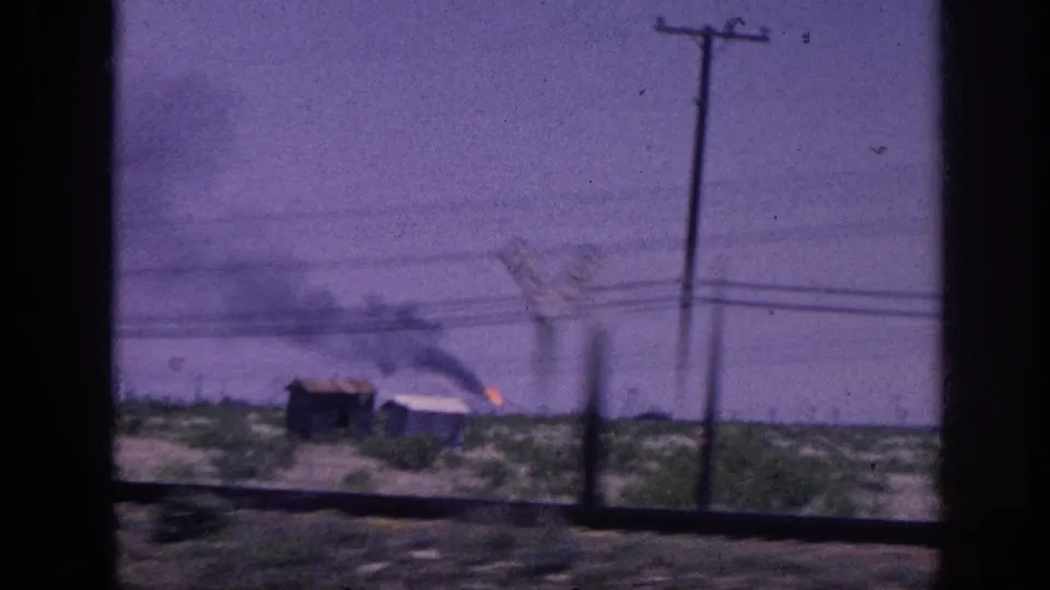 1963 Memphis, Tennessee area: vehicle window view of a small rural outbuilding with dark smoke and active fire, set in a flat open landscape with utility poles.