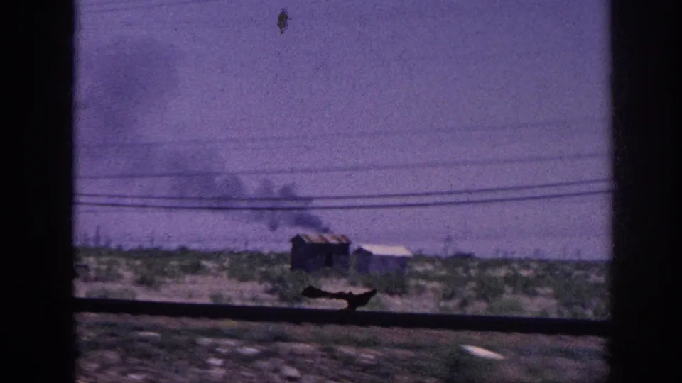 Through a moving vehicle's window, a small wooden outbuilding sends dark smoke skyward across flat rural terrain; 8mm footage metadata places this in the Memphis, Tennessee area, 1963.