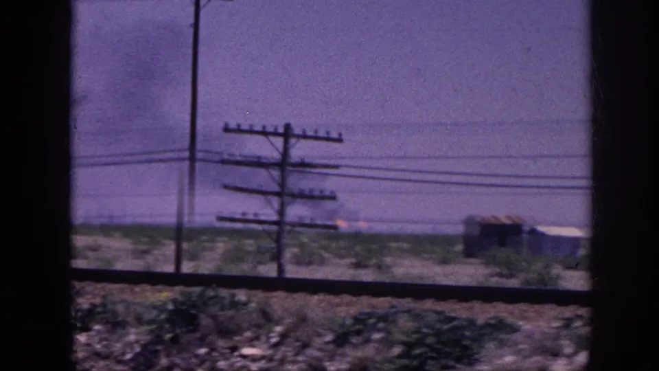 Mid-journey view from a moving vehicle showing flat rural terrain with utility poles and a faint smoke column at right; 8mm color footage consistent with early 1960s American South road travel.