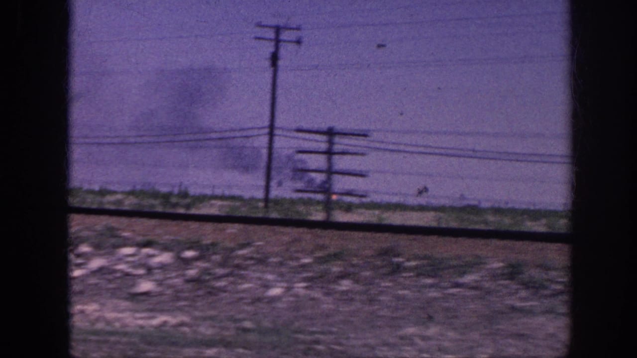 Moving-vehicle POV of flat rural scrubland with a telephone pole and a distant smoke plume rising under a purple-tinted sky.