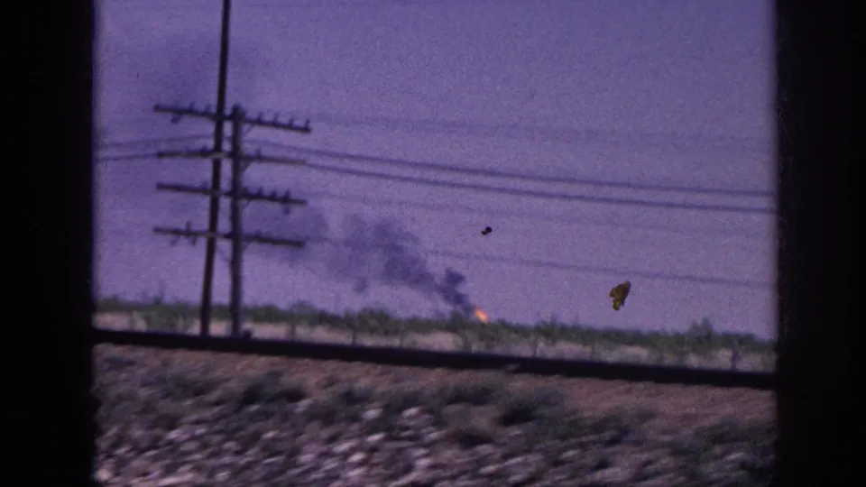 8mm driving shot of rural landscape with multiple telephone poles at left, a low fire burning near the horizon, and gravel debris on the road shoulder, likely Tennessee circa 1963.