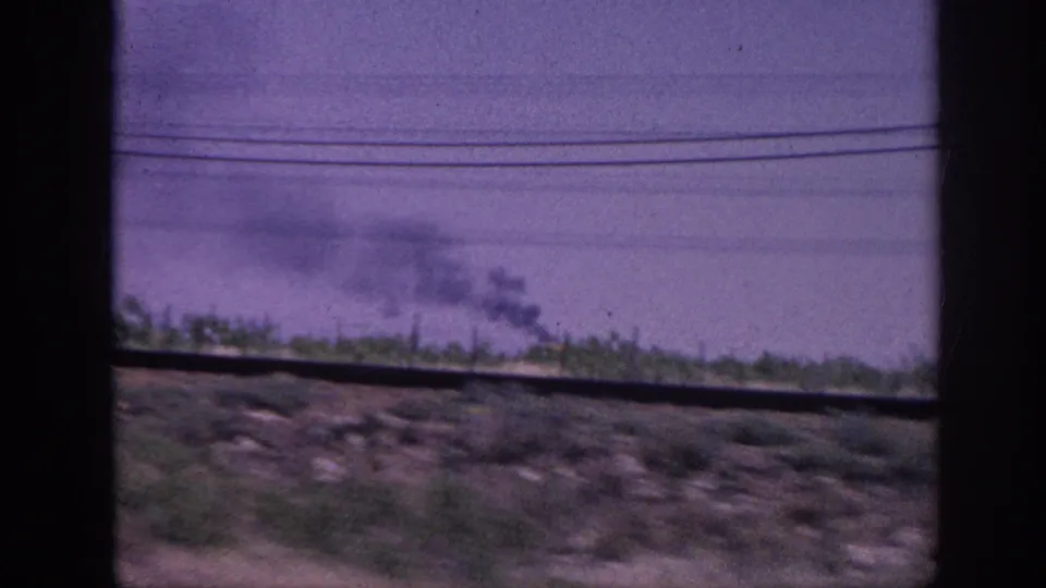 1963 8mm home movie: diffuse smoke on the horizon, multiple power lines, low roadside vegetation, distinctive purple color cast, shot from a moving vehicle.