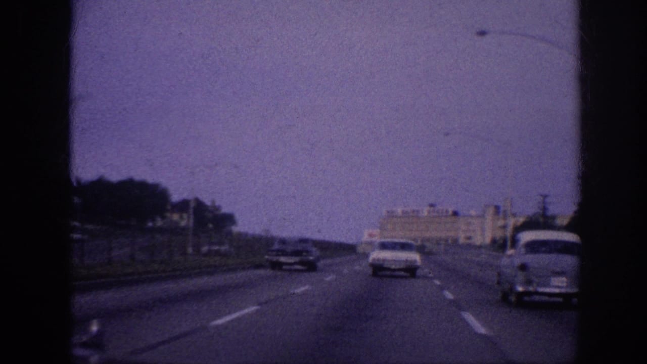 POV driving shot on a multi-lane 1960s American highway; two classic sedans ahead, large commercial building with sign in background, strong purple film tint.