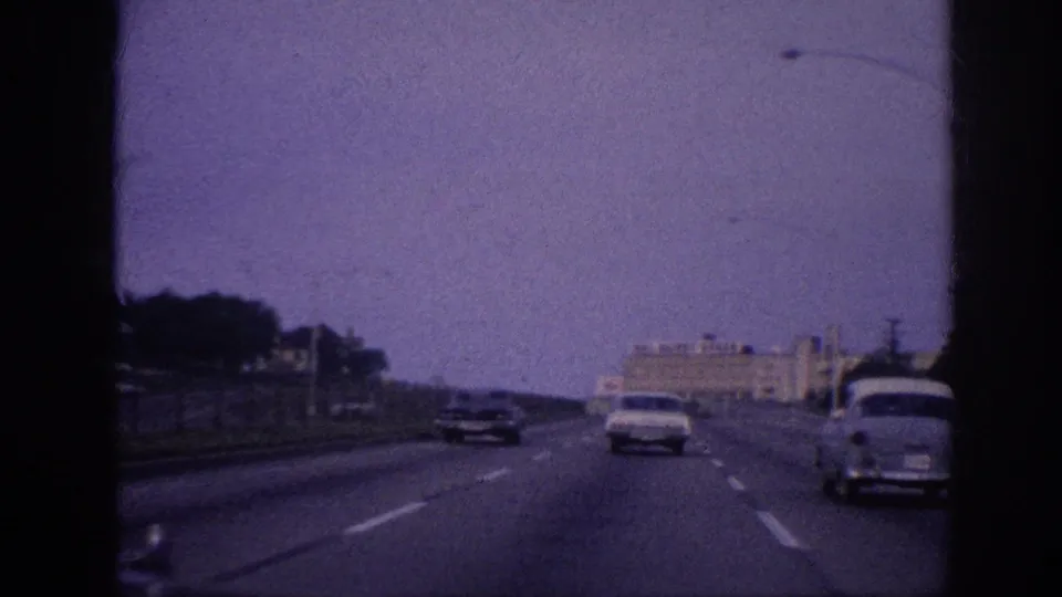 First-person windshield view on a Memphis, Tennessee highway circa 1963; early-1960s sedans ahead, Mos Bairo's Bread building visible in background, 8mm purple dye-fade.