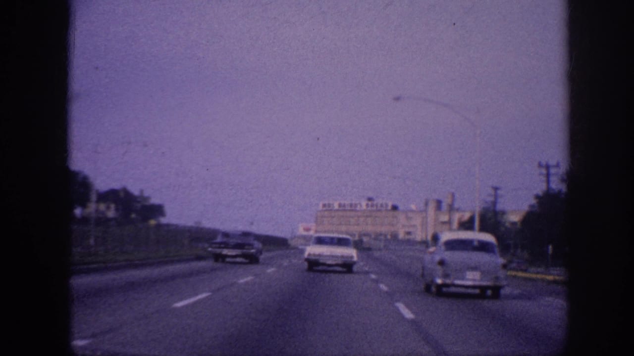 POV driving shot on 1960s multi-lane highway; white sedan ahead in center lane, Mos Bairo's Bread bakery building prominent right side, purple-tinted 8mm film grain.