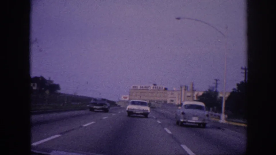 Windshield view on Memphis, Tennessee highway circa 1963; overhead streetlamp gantry spans the road, classic 1960s sedans ahead, Mos Bairo's Bread building right, 8mm purple tint.