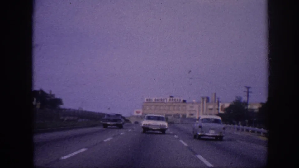 Windshield driving view on a Memphis, Tennessee highway circa 1963; light-colored classic sedan ahead, Mos Bairo's Bread building to the right, 8mm grain with pronounced purple dye cast.