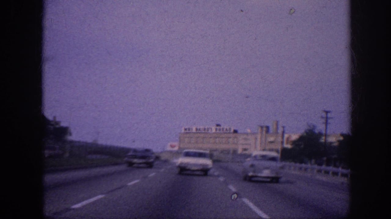 Windshield POV on a Memphis freeway, 1963; two early-1960s American sedans ahead; Mrs. Bairo's Bread industrial building visible in the background.
