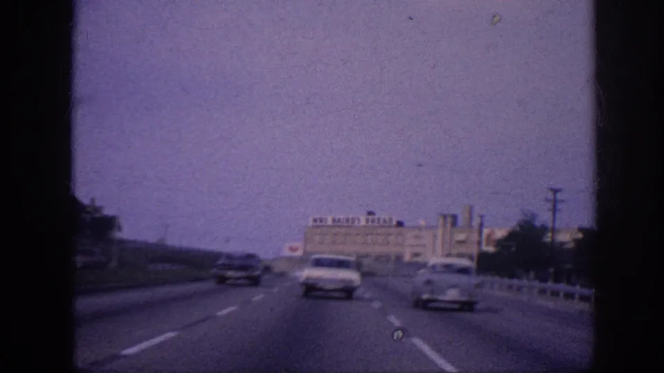 Windshield POV on a Memphis freeway, 1963; two early-1960s American sedans ahead; Mrs. Bairo's Bread industrial building visible in the background.