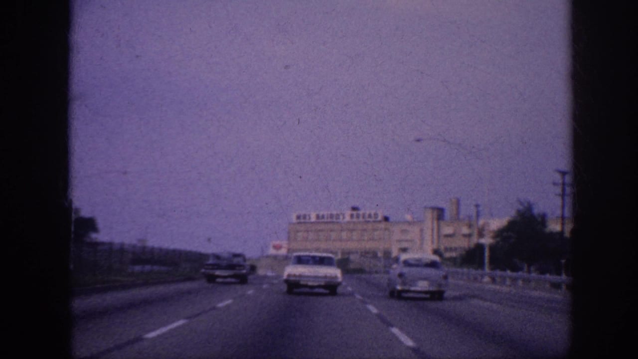 Freeway from moving vehicle; two early-1960s American cars ahead; industrial bakery building with rooftop sign more prominently in view.