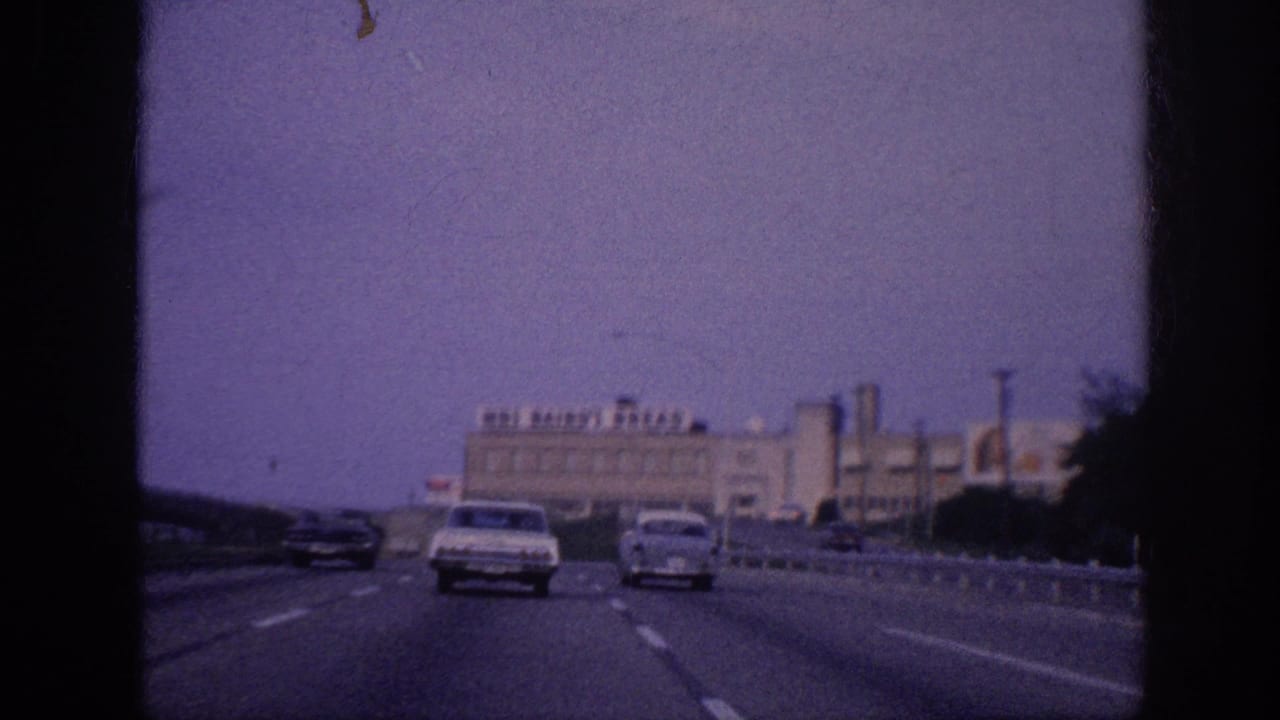 1963 Memphis road-trip footage shows mid-century cars traveling on a divided highway toward a large Mrs Baird's Bread factory building under a purple-shifted sky.