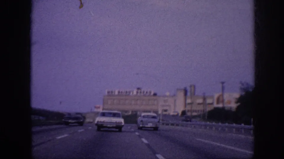 1963 Memphis road-trip footage shows mid-century cars traveling on a divided highway toward a large Mrs Baird's Bread factory building under a purple-shifted sky.
