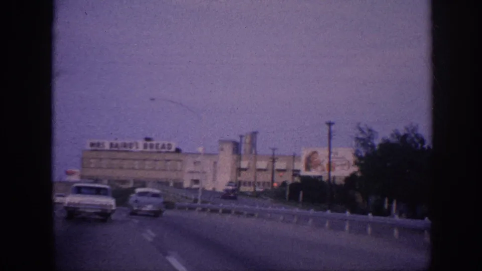 A moving-vehicle view in Memphis shows cars on the highway passing the large Mrs Baird's Bread building, with roadside poles and trees along the edge of the frame.