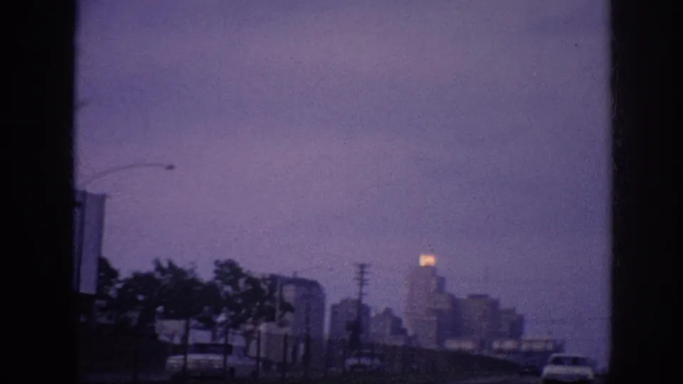 The later road-trip view turns toward a distant Memphis skyline at dusk, with dark roadside trees, utility poles, and a softly glowing building top visible through grainy 8mm color.