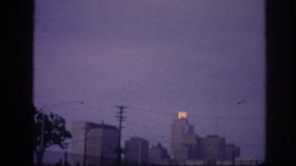 8mm home-movie frame from 1963, possibly Memphis, Tennessee — silhouetted downtown skyline at dusk, utility poles, and a glowing rooftop sign against a violet sky.