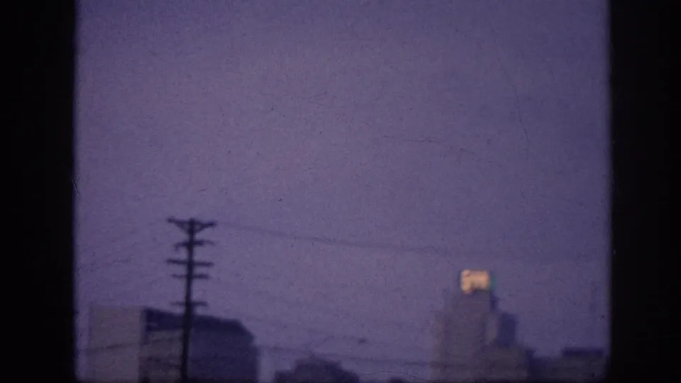 8mm frame at 0:02, possibly Memphis 1963 — utility pole crossarm centered, city skyline in silhouette, and a glowing rooftop sign on a right-side high-rise at dusk.
