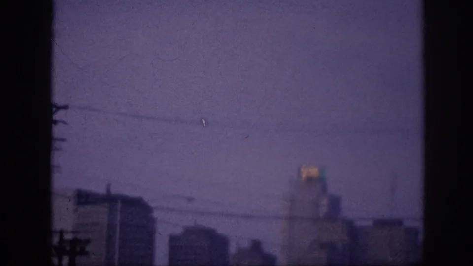 8mm frame at 0:03, possibly Memphis 1963 — downward-shifted skyline view, power lines crossing the lower foreground, and a lit rooftop sign under a violet sky.