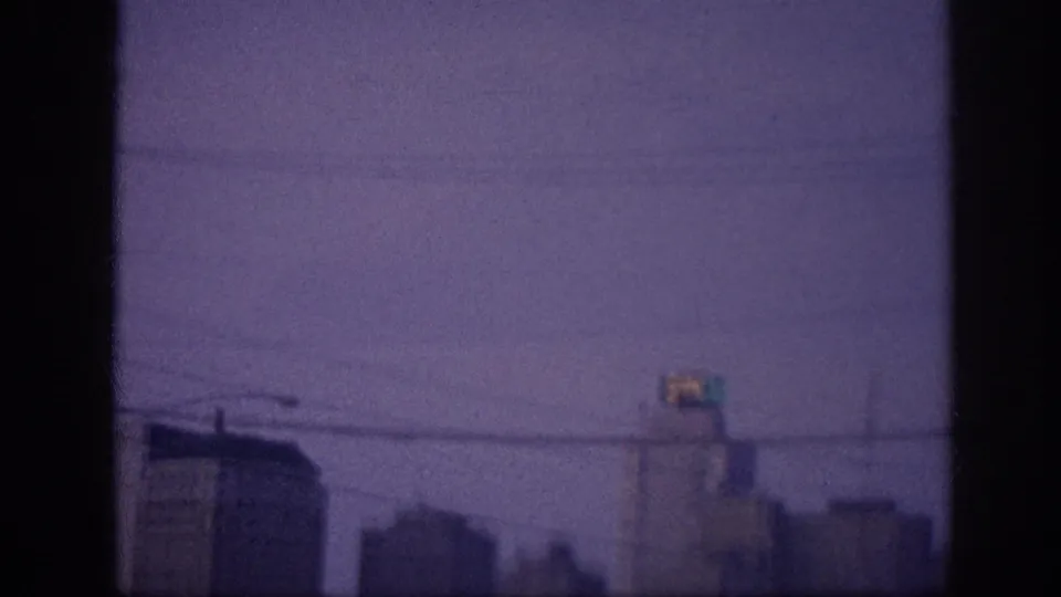 8mm frame at 0:05, possibly Memphis 1963 — rightward pan of dusk skyline with power lines and a color-shifted illuminated rooftop sign against a violet sky.