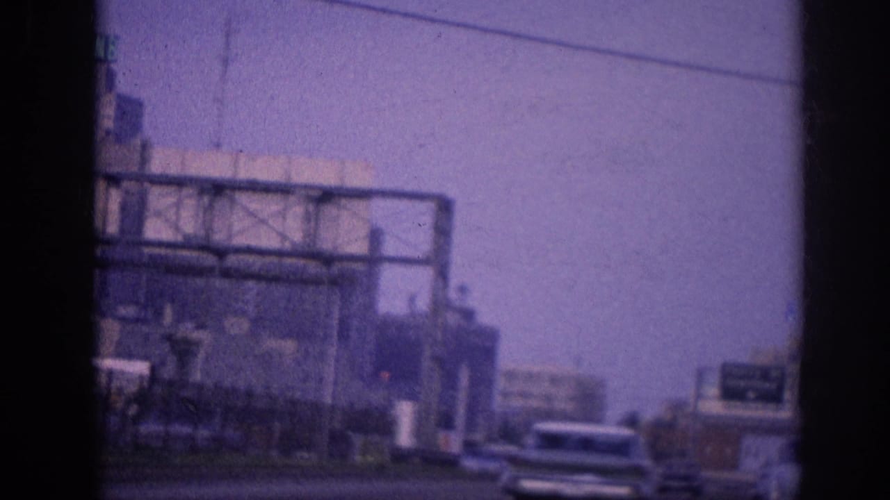 Moving-vehicle view of early-1960s American sedans on a multi-lane city highway under a hazy sky