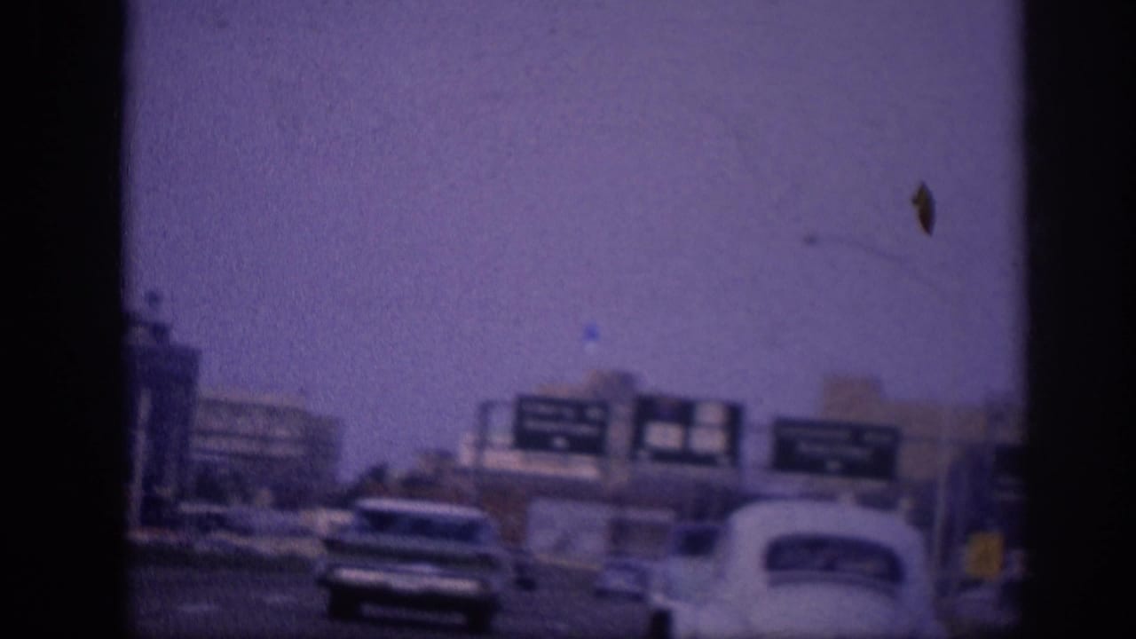 Road-level view of a large white American sedan on a city highway with green directional signs overhead and urban buildings behind