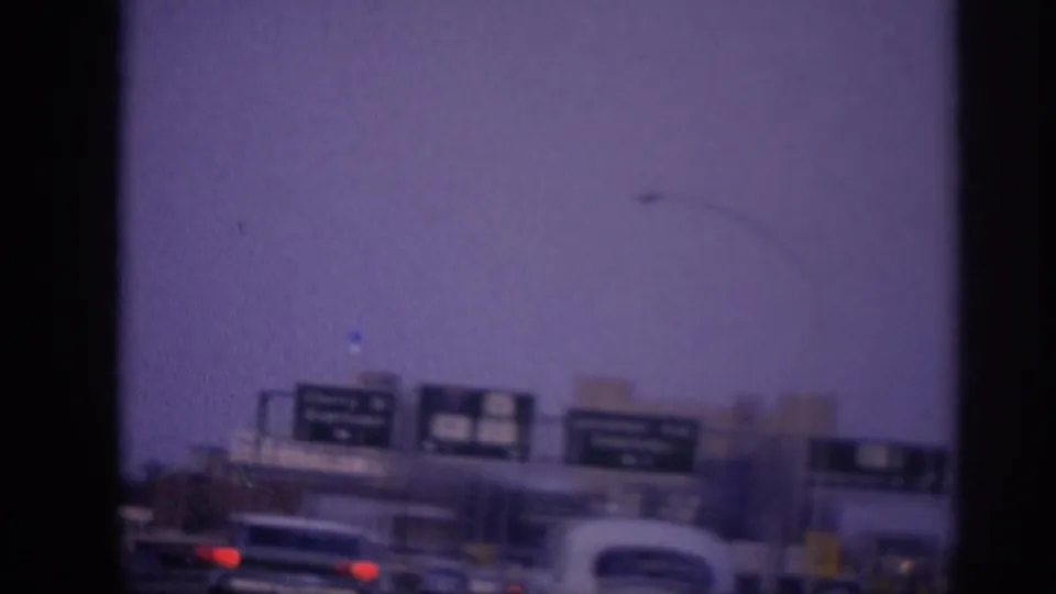 8mm film frame showing a multi-lane city highway in Memphis circa 1963, with early-1960s American sedans, red taillights visible ahead, a street lamp, and multiple green directional sign panels overhead.