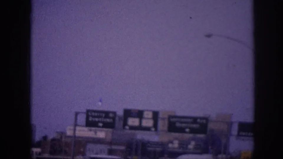 8mm home movie frame showing multiple green directional highway sign panels spanning a Memphis city freeway, early-1960s American cars in traffic, urban buildings, and a street lamp at upper right, circa 1963.