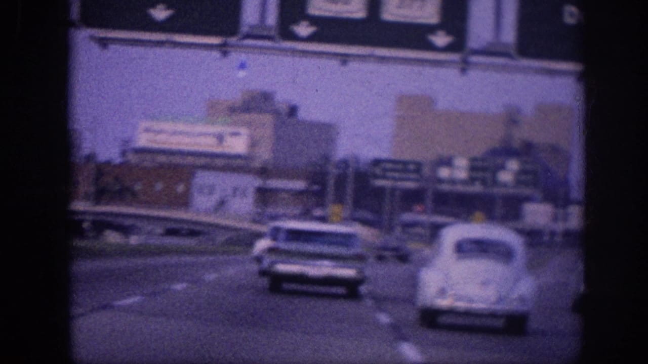8mm film frame circa 1963 Memphis area: white Volkswagen Beetle and American sedan on a multi-lane expressway, with overhead highway signs and commercial structures in background.
