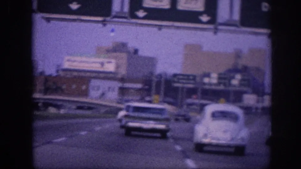 8mm film frame circa 1963 Memphis area: white Volkswagen Beetle and American sedan on a multi-lane expressway, with overhead highway signs and commercial structures in background.