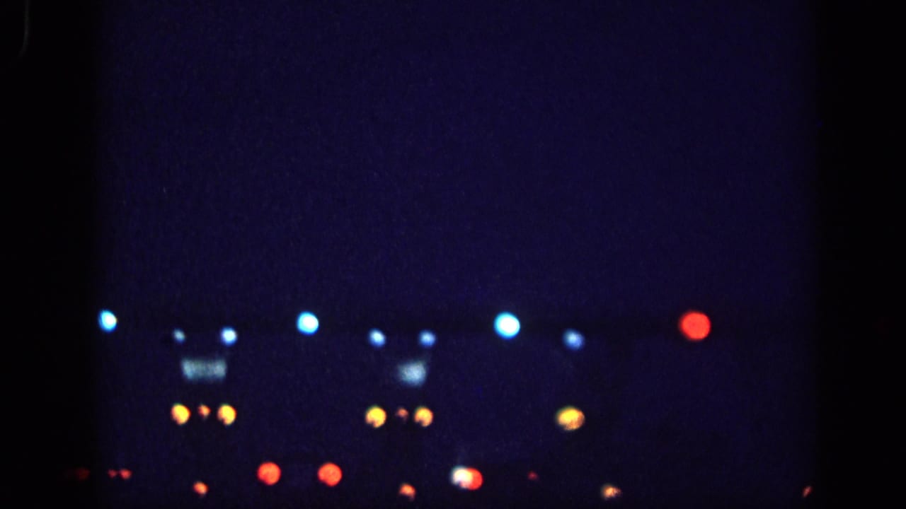 8mm home-movie frame circa 1963, possibly Memphis, Tennessee: scattered blue, amber, and red bokeh lights at night, with an amber cluster suggesting a rectangular illuminated sign or structure.